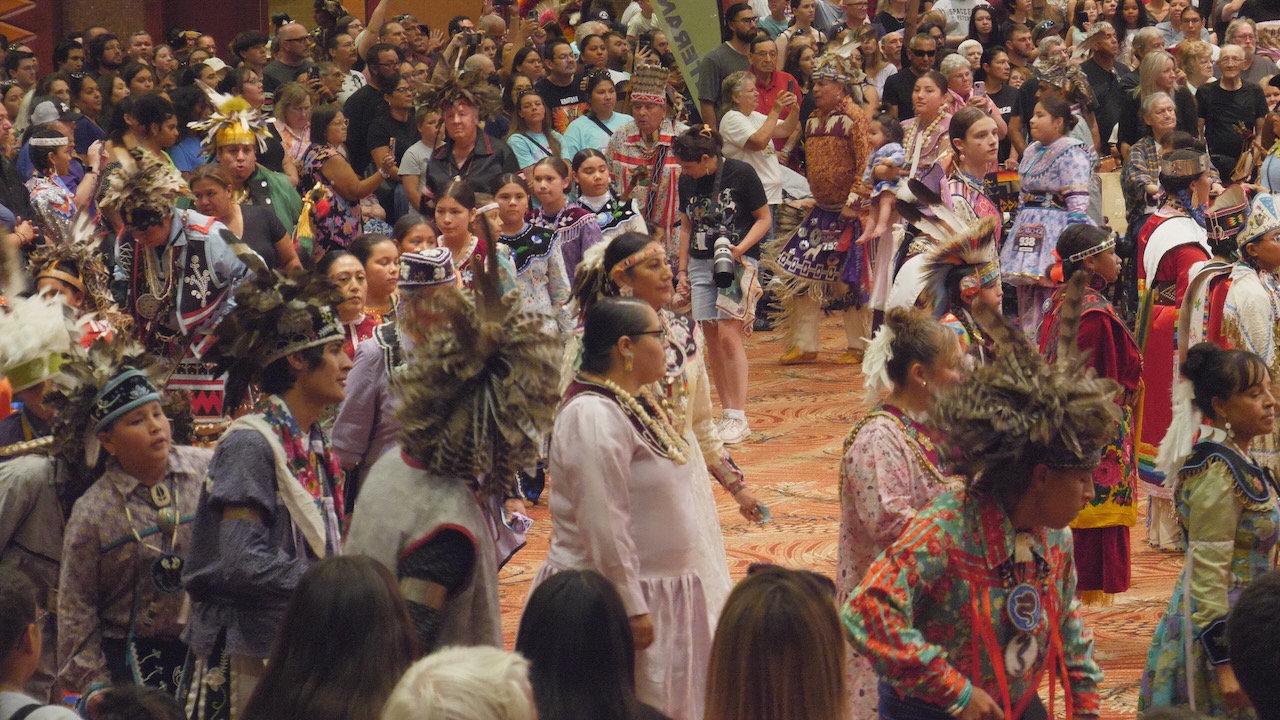 A circle of Haudenosaunee dancers of all genders and ages in traditional regalia dances, surrounded by onlookers, at the Salamanca Powwow (July 20, 2024)